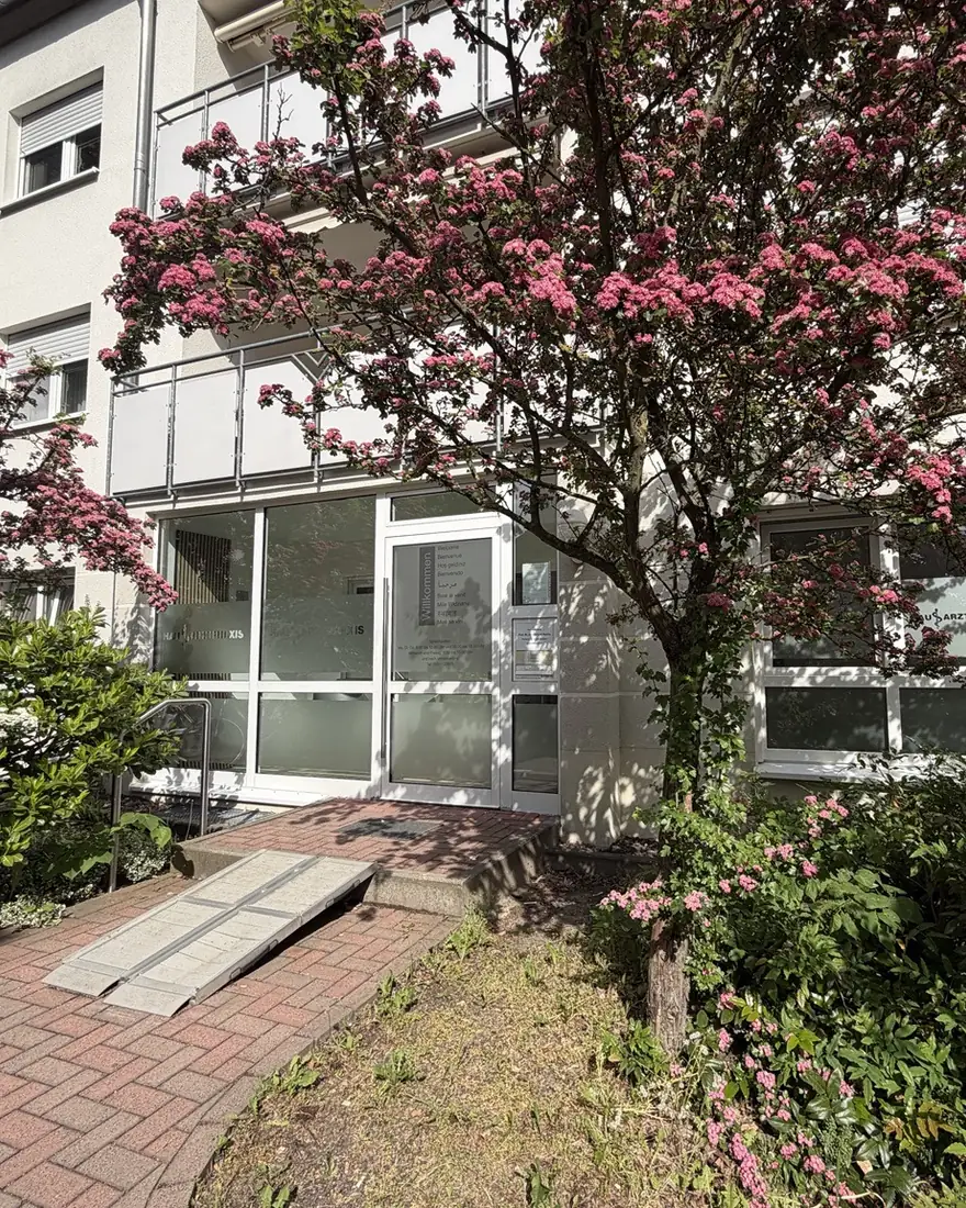 Eingang der Hausarztpraxis in der Kleberstraße 26: Glastür mit Rampe unter einem blühenden Baum